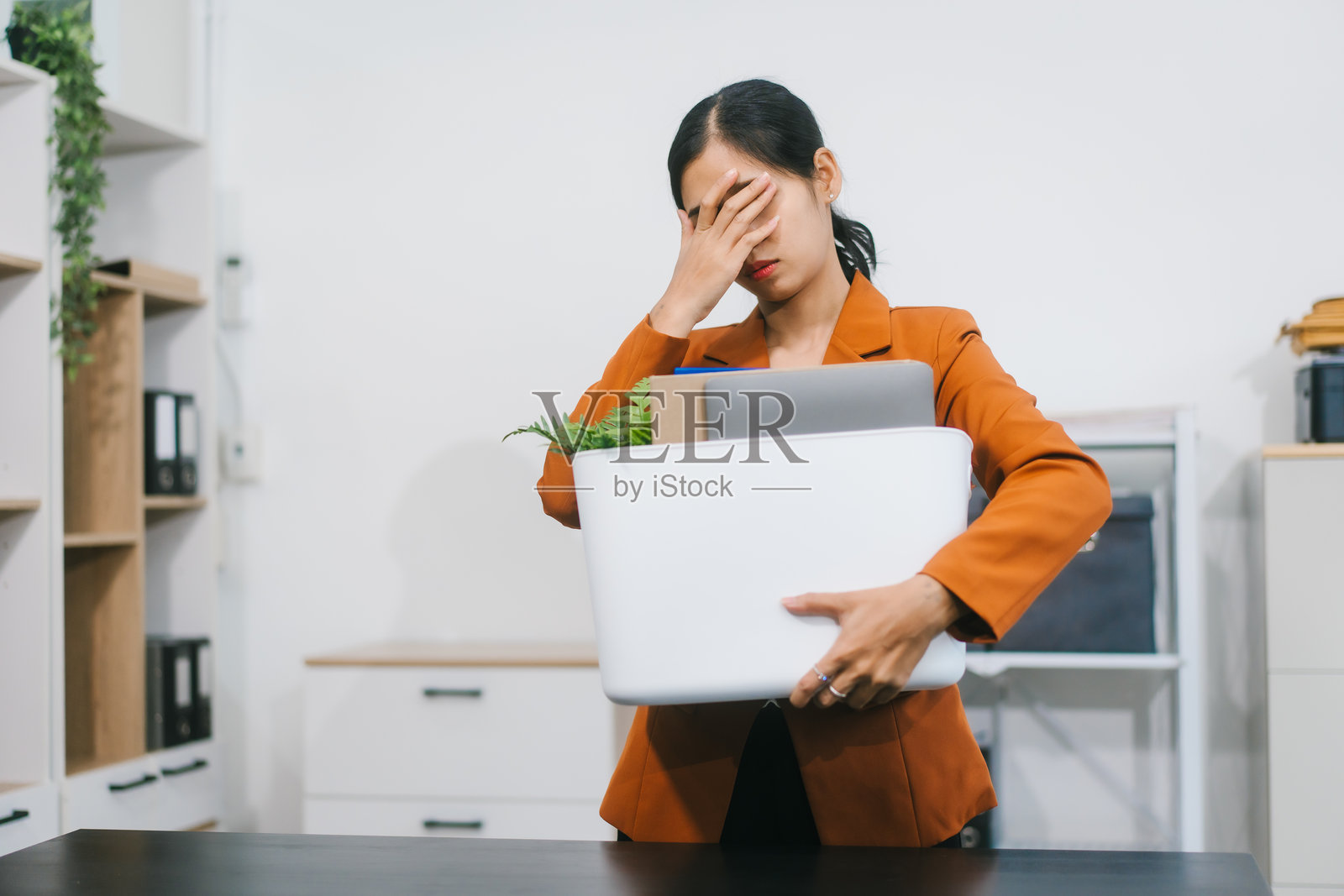 一位亚洲女性独自坐在办公室里,显得压力重重、沮丧不已,身边放着一箱个人物品,象征着失业、辞职或工作中的挑战。照片摄影图片