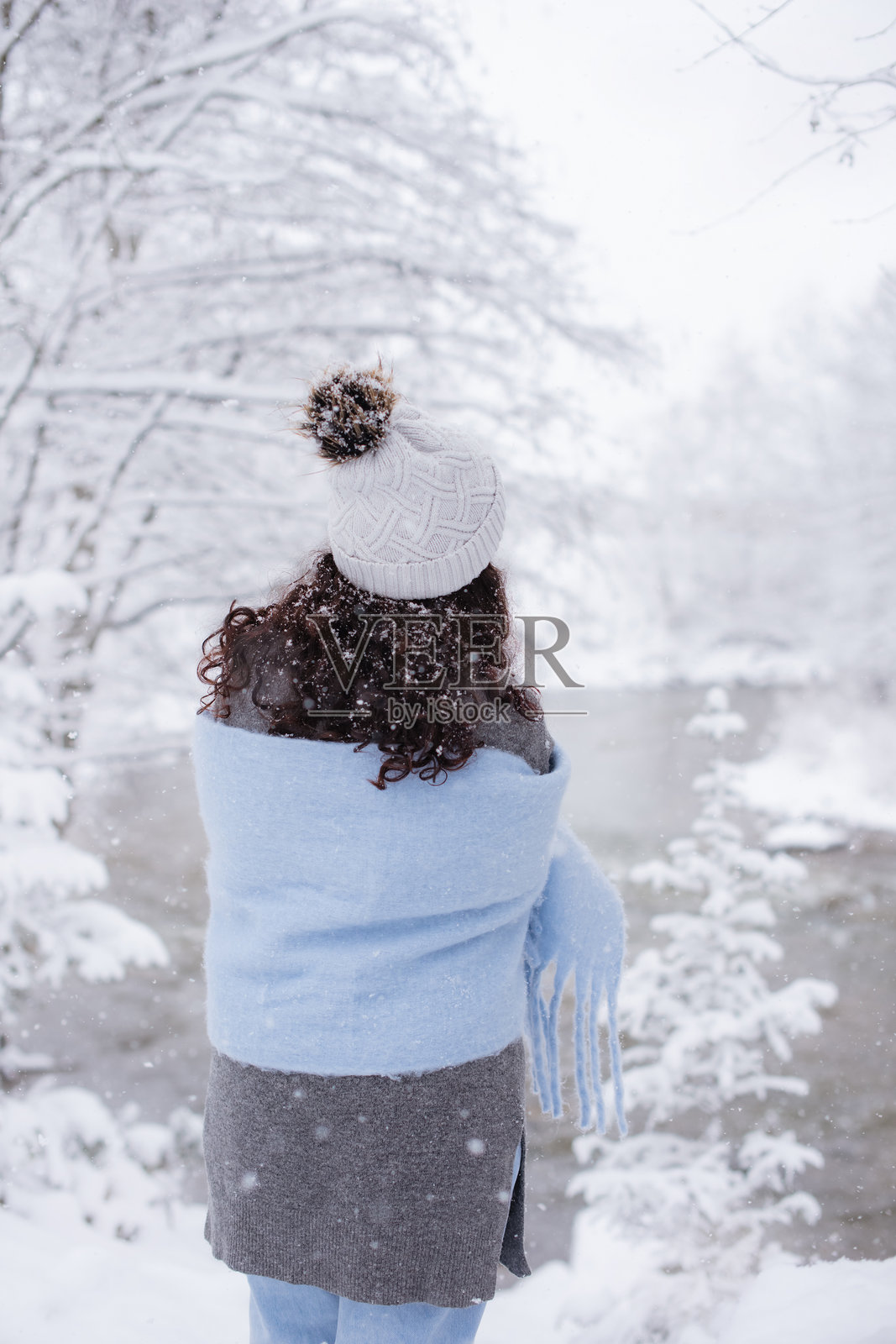 身披蓝色围巾的女子走在雪林中,站在河岸边照片摄影图片