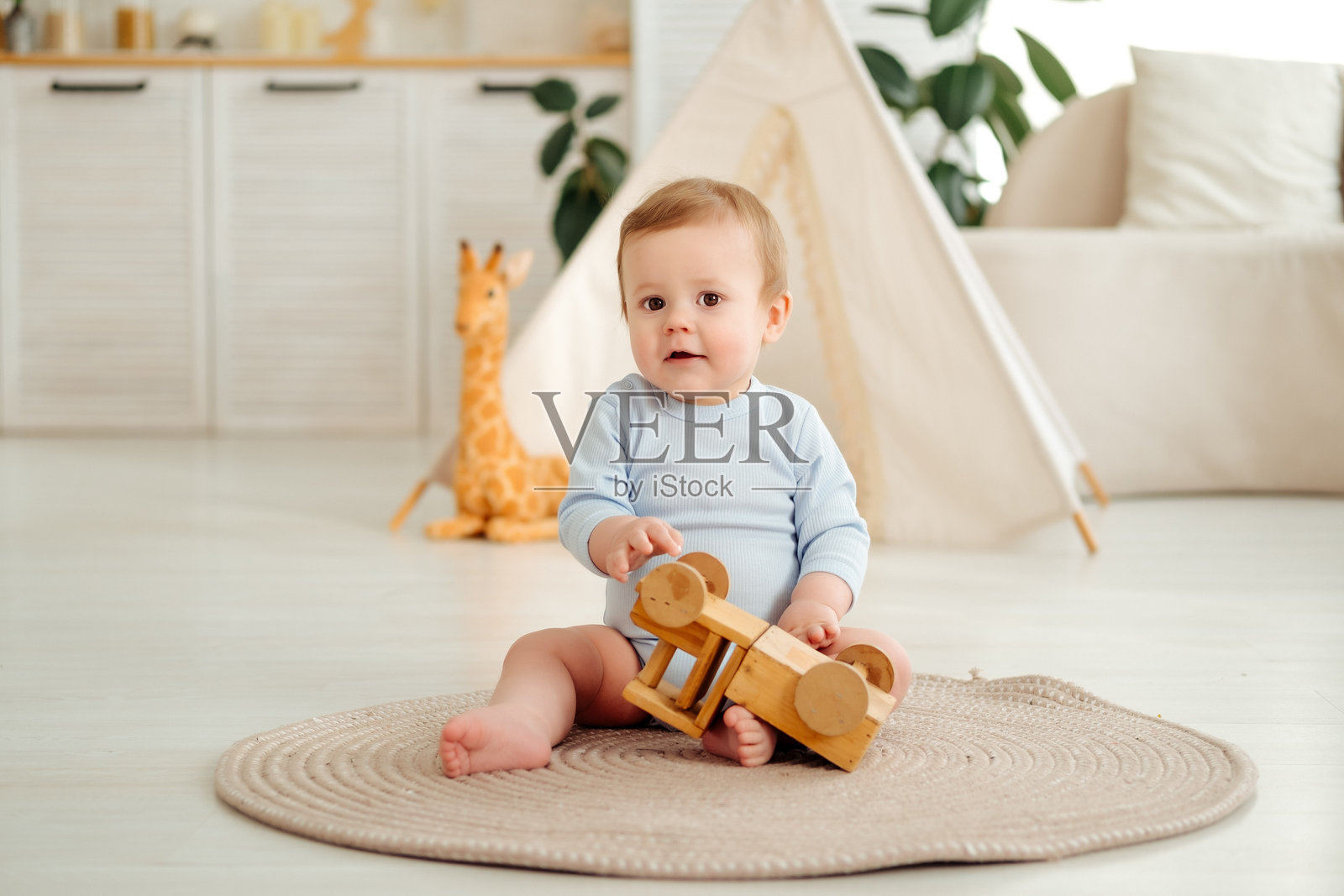 一个快乐的微笑孩子在家里的儿童房里,在印第安帐篷旁边玩着木制玩具车,一个小婴儿穿着蓝色连体衣坐着大笑照片摄影图片