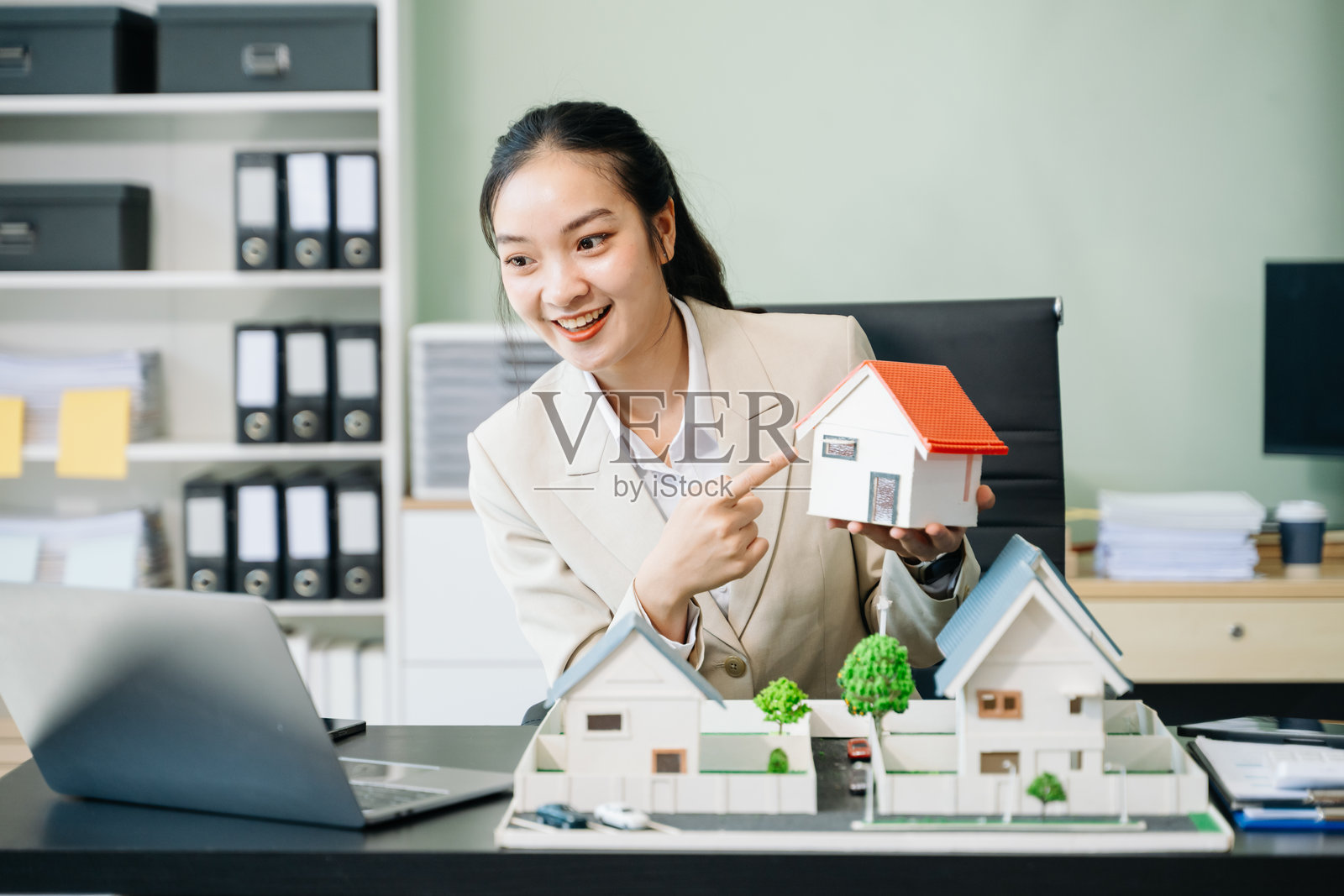 年轻的房地产经纪人坐在桌旁,使用笔记本电脑和平板电脑工作,旁边放着一个小房子。照片摄影图片