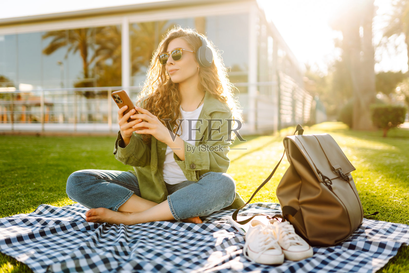 年轻女子戴着无线耳机,在阳光明媚的公园里,周围环绕着棕榈树,她一边坐着一边玩手机。照片摄影图片