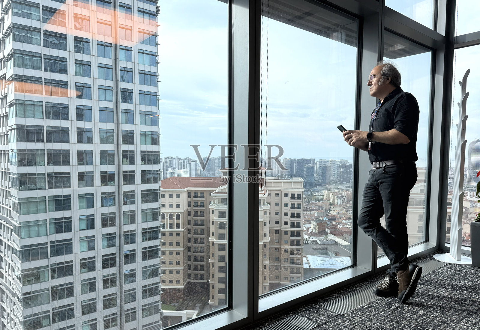 一位深思熟虑的商人站在办公室里,看着窗外的大都市,同时使用智能手机。这位富有魅力的数字企业家正在为电子商务初创公司规划投资策略。照片摄影图片