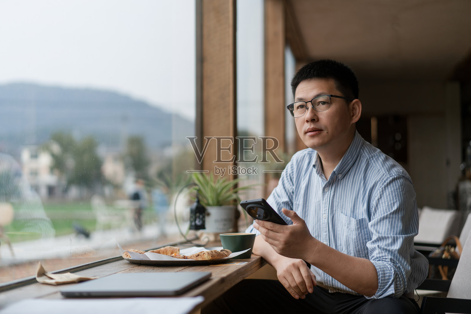 戴眼镜男子窗边用手机看窗外风景照片摄影图片