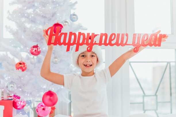 滑稽的funky男孩正举起红色的新年祝福语,坐在圣诞树旁的白色床毯上。图片 滑稽的funky男孩正举起红色的新年祝福语,坐在圣诞树旁的白色床毯上。图片下载