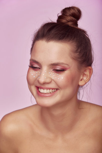 时尚创意化妆。年轻女子的面部表情与干净的新鲜的皮肤和明亮的妆与莱茵石雀斑在她的脸上摆姿势在明亮的粉红色背景图片 时尚创意化妆。年轻女子的面部表情与干净的新鲜的皮肤和明亮的妆与莱茵石雀斑在她的脸上摆姿势在明亮的粉红色背景图片下载