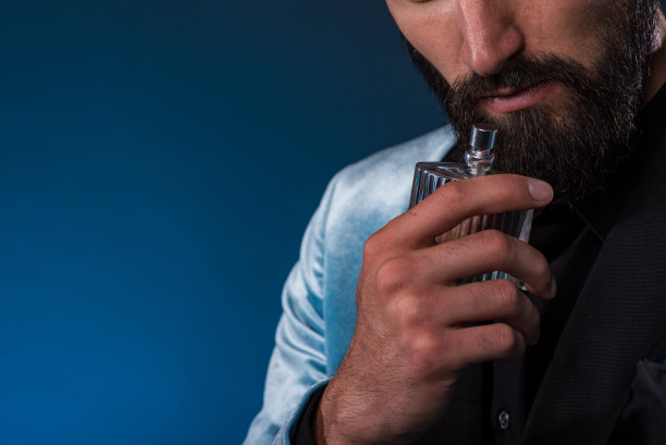 有男子气概的留着胡子的年轻人拿着一瓶普通的香水。男士香水、香水、化妆品。男性香水。大胡子男人拿着一瓶香水。图片 有男子气概的留着胡子的年轻人拿着一瓶普通的香水。男士香水、香水、化妆品。男性香水。大胡子男人拿着一瓶香水。图片下载