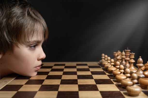 孩子下棋。男孩看木制的白色棋子在木板上。爱好、领导能力、智力和成功的概念。图片 孩子下棋。男孩看木制的白色棋子在木板上。爱好、领导能力、智力和成功的概念。图片下载
