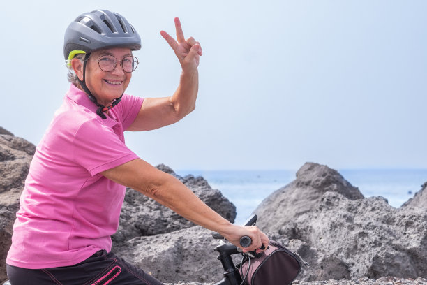积极快乐的高级妇女与头盔跑在她的电动自行车沿海看着相机胜利的标志。自由和假期的概念图片 积极快乐的高级妇女与头盔跑在她的电动自行车沿海看着相机胜利的标志。自由和假期的概念图片下载