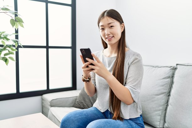 年轻的亚洲女人坐在客厅的沙发上使用智能手机图片 年轻的亚洲女人坐在客厅的沙发上使用智能手机图片下载