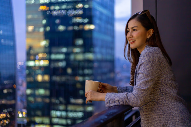 年轻的亚洲女子在阳台上欣赏夜景,一边看着城市摩天大楼的城市景观,一边喝着一杯热咖啡,为住房和现代生活方式的概念服务图片 年轻的亚洲女子在阳台上欣赏夜景,一边看着城市摩天大楼的城市景观,一边喝着一杯热咖啡,为住房和现代生活方式的概念服务图片下载