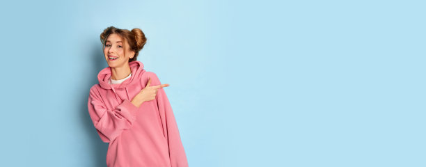 年轻的正面女孩的肖像在粉红色卫衣摆姿势,指着蓝色的工作室背景兴奋。好消息。摩天观景轮图片 年轻的正面女孩的肖像在粉红色卫衣摆姿势,指着蓝色的工作室背景兴奋。好消息。摩天观景轮图片下载