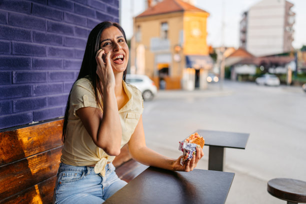 年轻女子在户外快餐店坐着,一边吃三明治一边打电话图片 年轻女子在户外快餐店坐着,一边吃三明治一边打电话图片下载