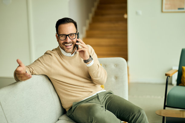 快乐的男人坐在沙发上,在现代客厅里打电话,面带微笑,神情投入图片 快乐的男人坐在沙发上,在现代客厅里打电话,面带微笑,神情投入图片下载