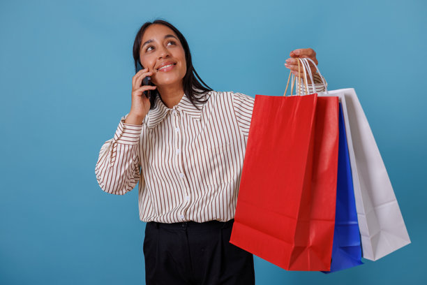 快乐的女人拎着购物袋,一边打电话,在蓝色的背景下享受购物的乐趣。图片 快乐的女人拎着购物袋,一边打电话,在蓝色的背景下享受购物的乐趣。图片下载