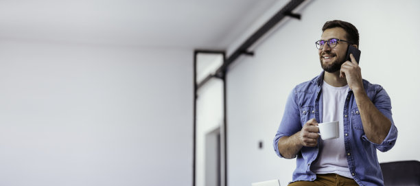 英俊的留着胡子的男人靠在办公桌上,一边喝咖啡一边打电话。图片 英俊的留着胡子的男人靠在办公桌上,一边喝咖啡一边打电话。图片下载