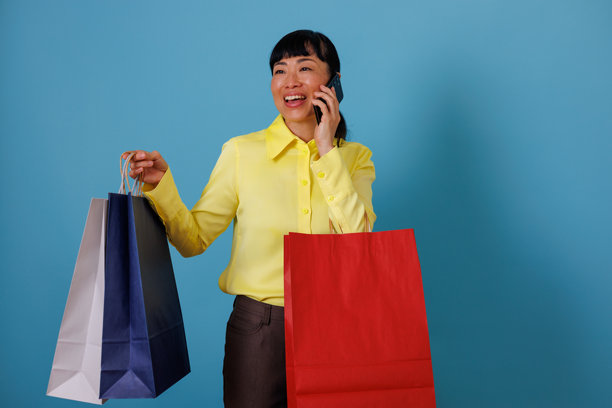 快乐的女人在蓝色的背景前购物,一边打电话,手里拿着装满商品的纸袋。图片 快乐的女人在蓝色的背景前购物,一边打电话,手里拿着装满商品的纸袋。图片下载