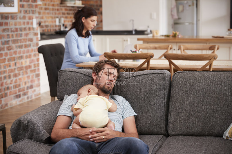 疲惫的父亲和婴儿在沙发上睡觉,而母亲在工作正版图片素材