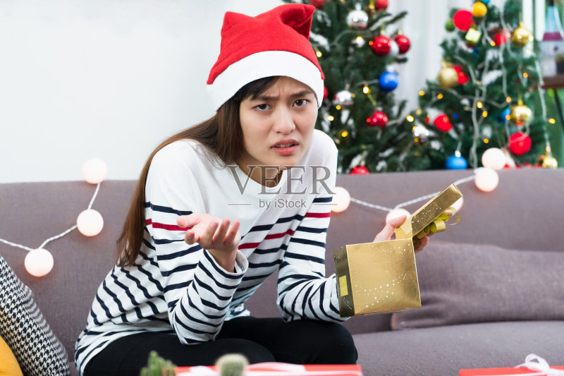 亚洲女人心烦意乱的时候在沙发上打开金色的圣诞礼盒,无聊的圣诞派对礼物。正版图片素材