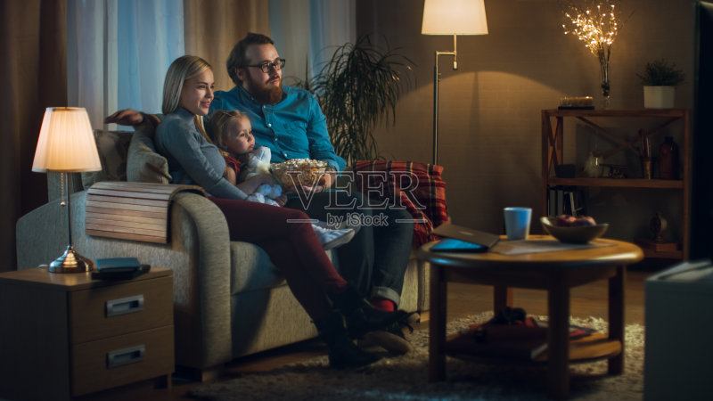 父亲,母亲和小女孩看电视的可能性不大。他们坐在舒适的客厅里的沙发上吃着爆米花。这是晚上。正版图片素材