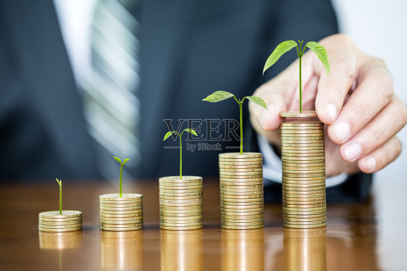 商人手里拿着一棵长着的钱币,把钱币堆成一个图形排列在木桌上,说明了金钱生长和存钱的概念正版图片素材