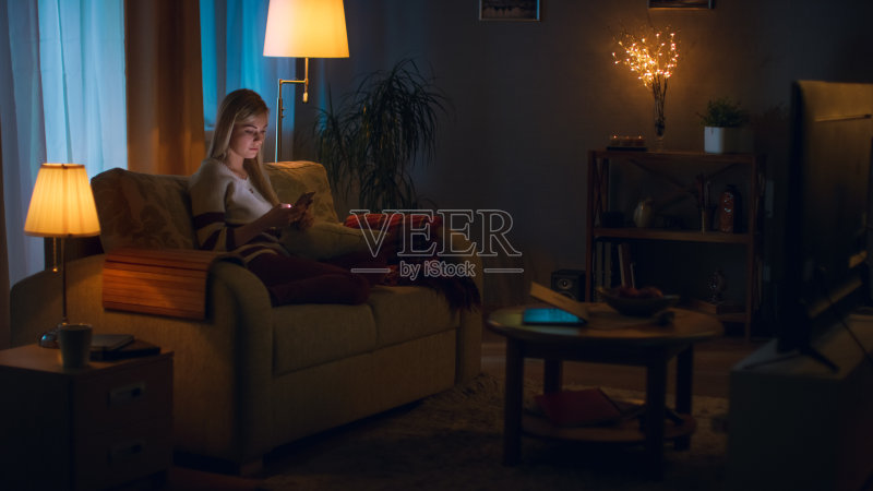 在晚上美丽的年轻女人放松在沙发上在她舒适的客厅。她使用智能手机。正版图片素材