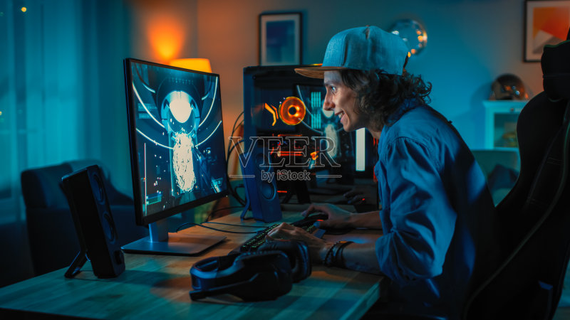 专业玩家在他强大的个人电脑上玩第一人称射击在线视频游戏。房间和电脑都有彩色霓虹灯。年轻人戴着一顶帽子。在家度过一个舒适的夜晚。正版图片素材