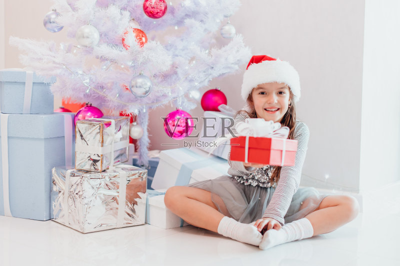 微笑的圣诞老人坐在冷杉树下,把包裹好的圣诞礼物递给镜头。正版图片素材