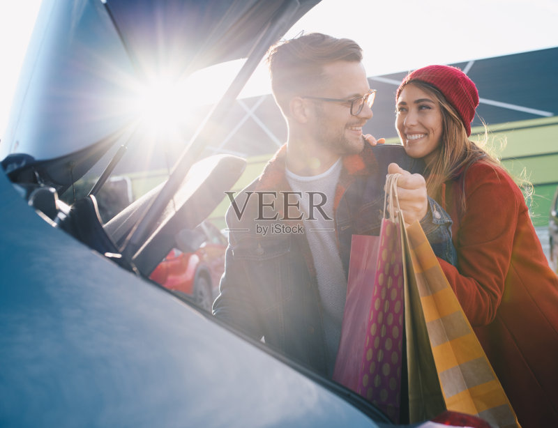 一对相爱的年轻情侣在购物中心前的停车场把购物袋塞进了汽车后备箱。购物后,他们开心地微笑着。正版图片素材