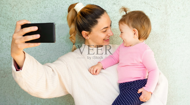 母女用智能手机自拍——妈妈抱着宝宝,开心地对着镜头摆姿势——母亲的生活方式正版图片素材