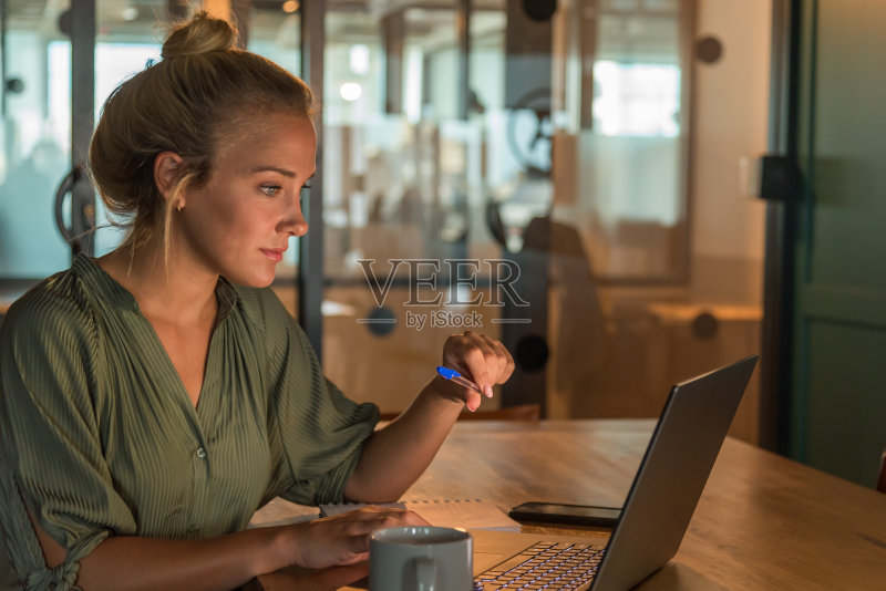 年轻的金发女人在她昏暗的笔记本前工作。桌子上有一个杯子,一个笔记本和一个手机正版图片素材