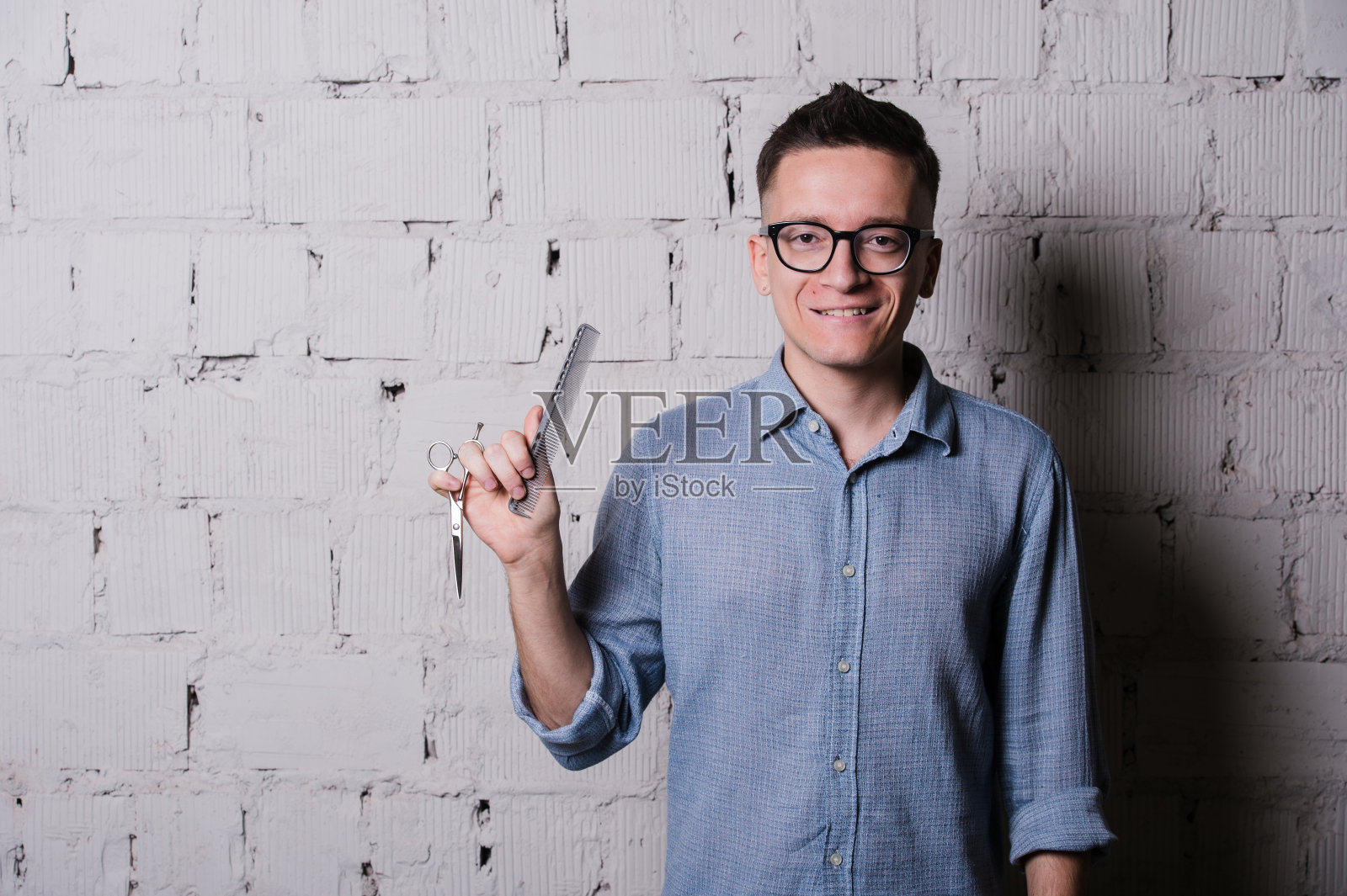 英俊的年轻男性理发师戴着眼镜微笑着，摆着剪刀的姿势照片摄影图片