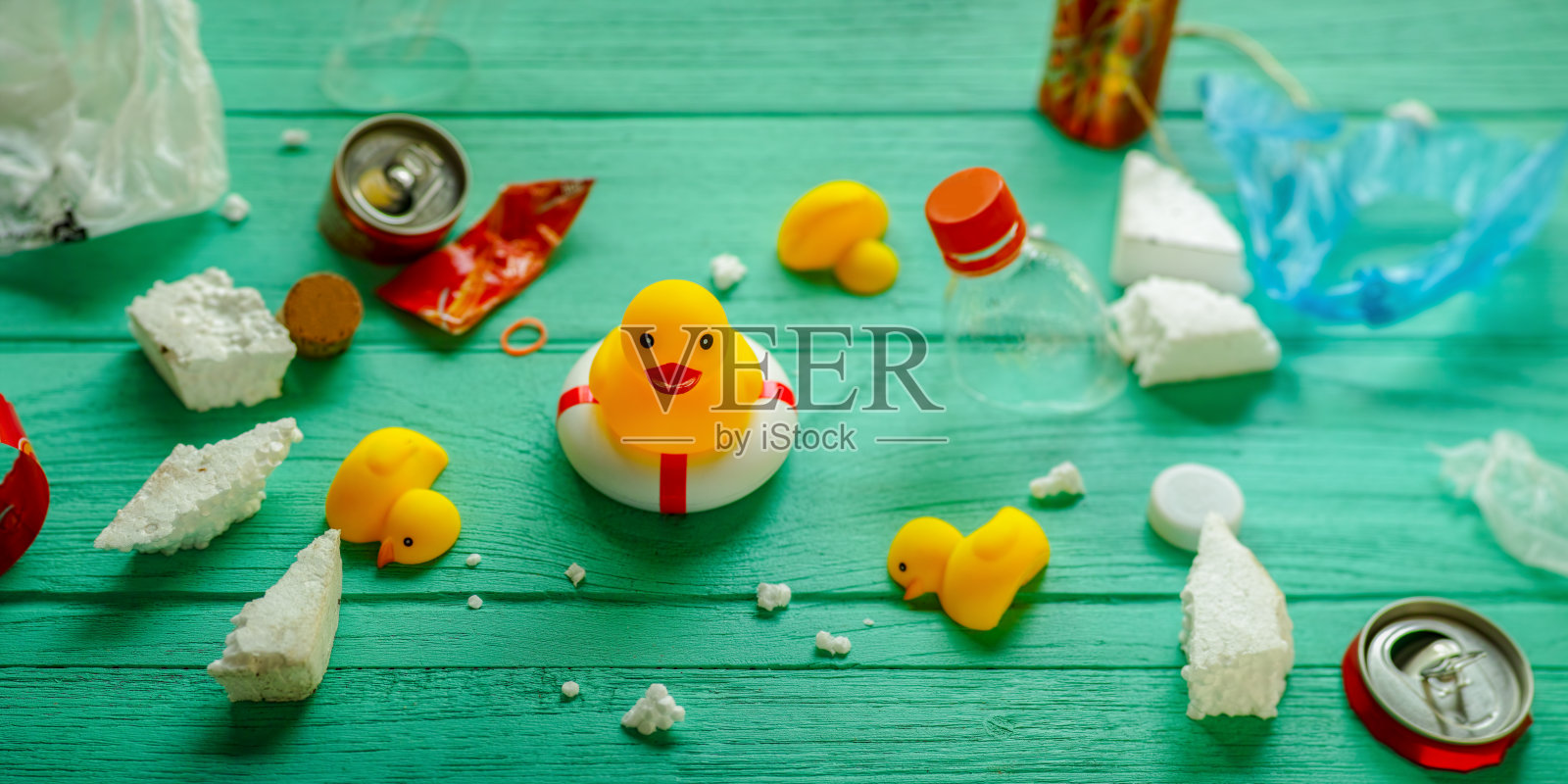 一只黄色的橡皮鸭站在一个充气式救生圈上，周围是漂浮的污染垃圾，还有三只死鸭放在一块绿松石色的木板上。照片摄影图片
