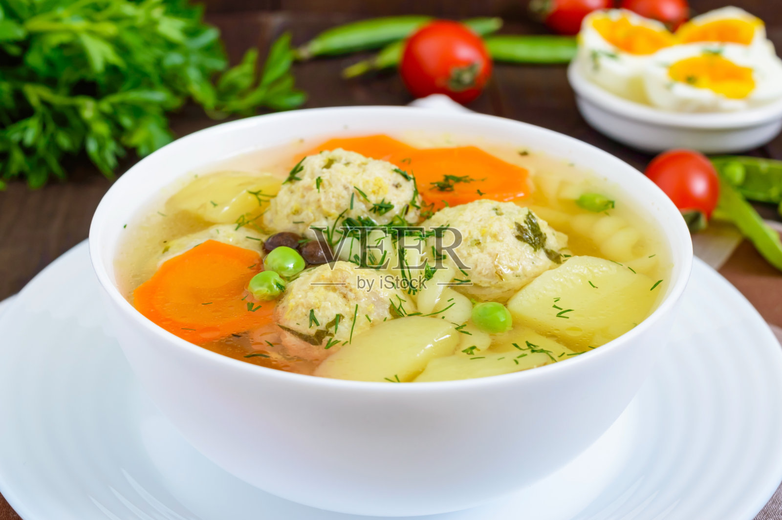 在深色的木桌上，配以肉丸、蔬菜、意大利面和煮鸡蛋的清淡汤。有用的午餐。照片摄影图片