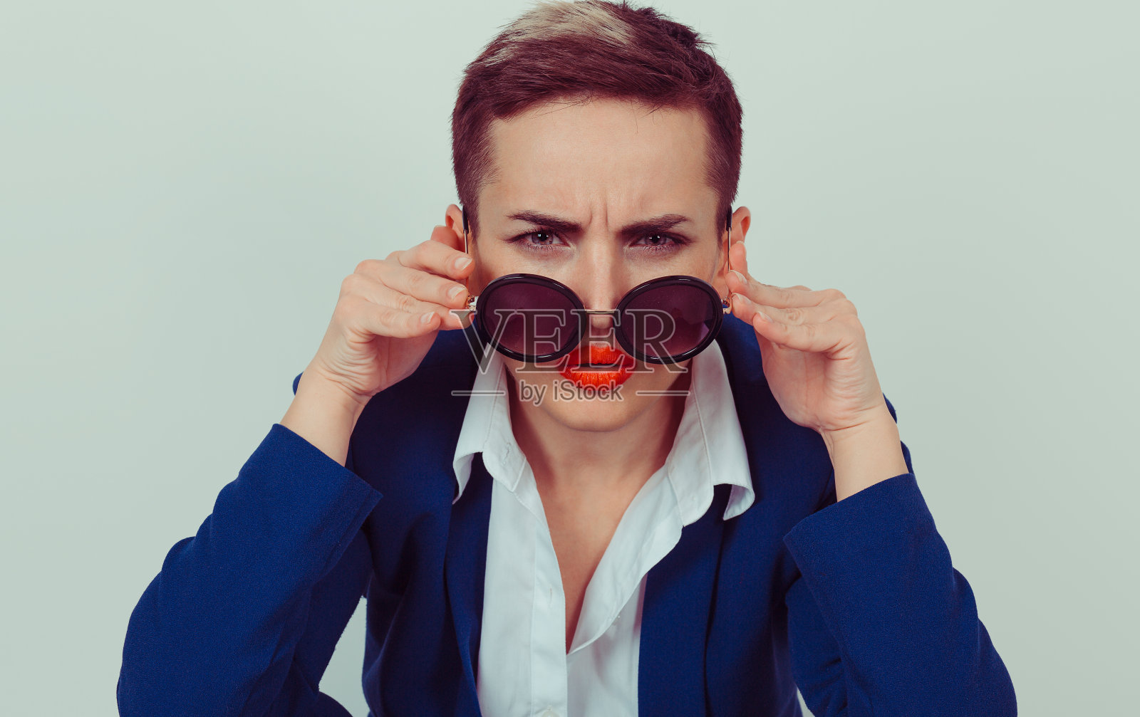 大头照严肃生气的泼妇妻子拿着太阳镜向下怀疑地看着你孤立的绿黄墙背景，蓝衬衫。人脸表情肢体语言，态度，感知照片摄影图片