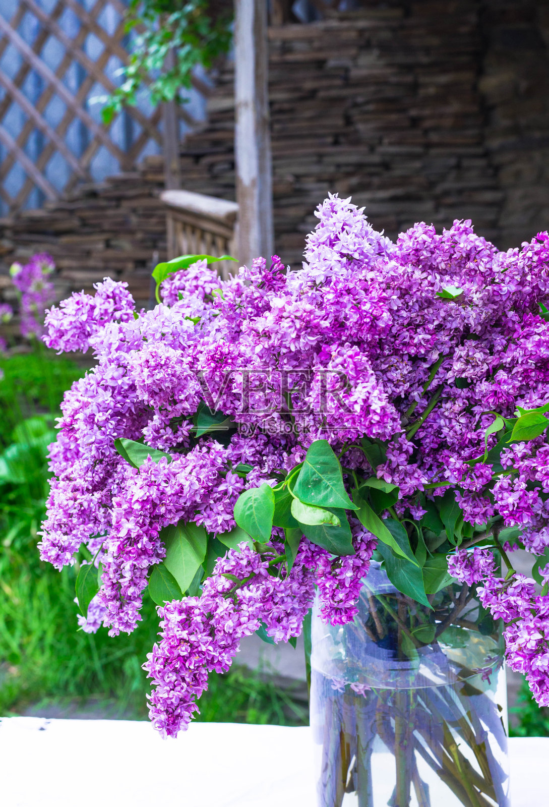 白色的复古花边桌布上，灰色的石墙上，花瓶里的丁香花束的美丽构图。照片摄影图片