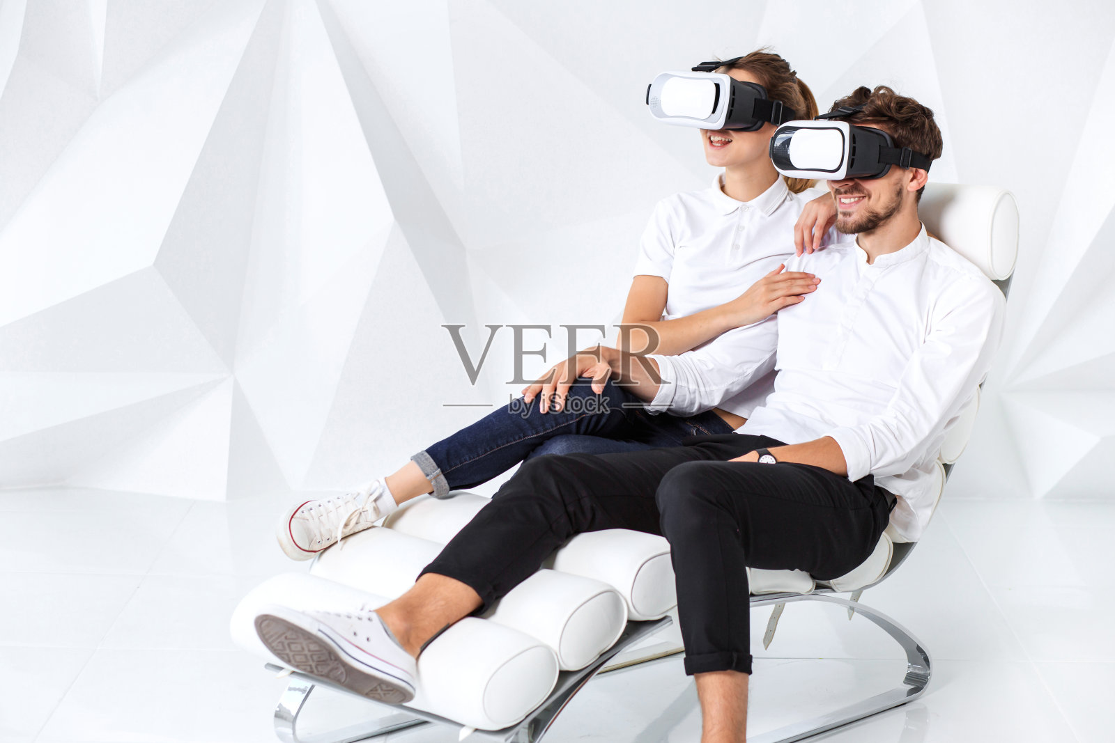 一对年轻夫妇戴着虚拟现实耳机坐在白色的椅子上，房间的墙壁和地板都是白色的照片摄影图片