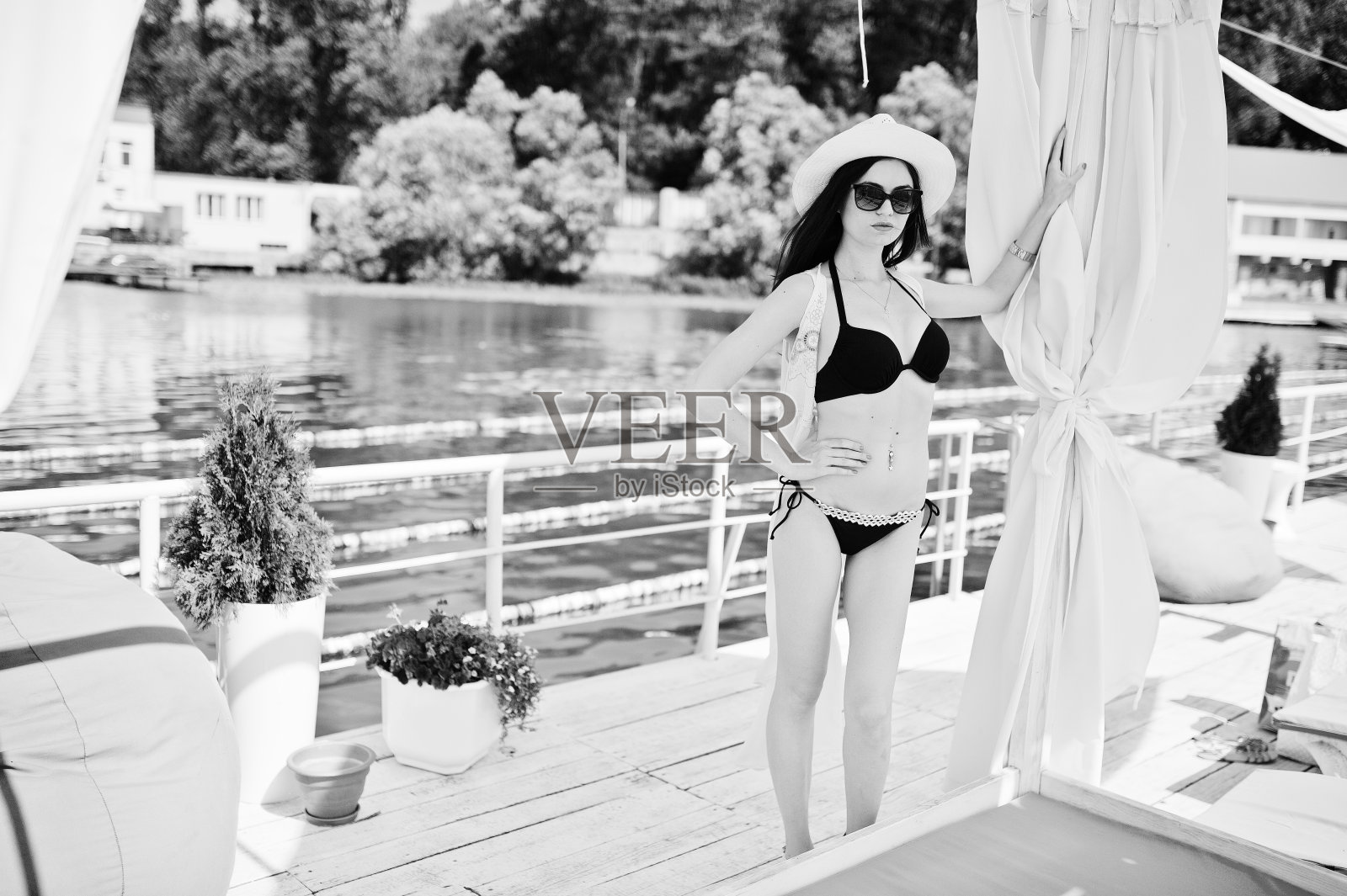 美丽的年轻女子在黑色比基尼泳装和透明斗篷摆姿势旁边的露台上戴着太阳镜。黑白照片。照片摄影图片
