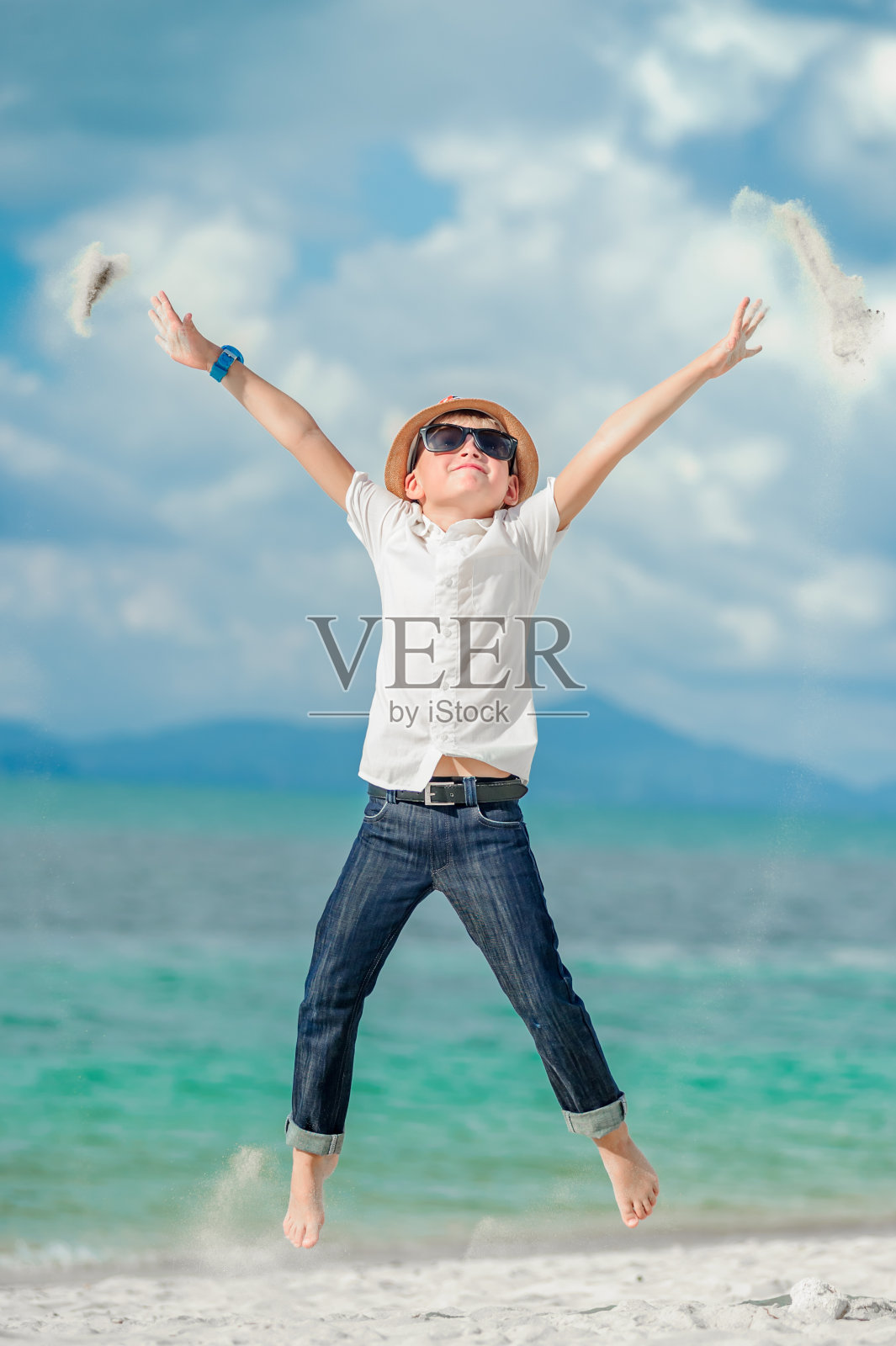 完整的生长肖像在一个热带海滩:英俊英俊的8岁男孩在固体明亮的修身合身衬衫，深色裤子太阳镜和软呢帽，赤脚在沙子上，跳跃，悬挂在空中照片摄影图片