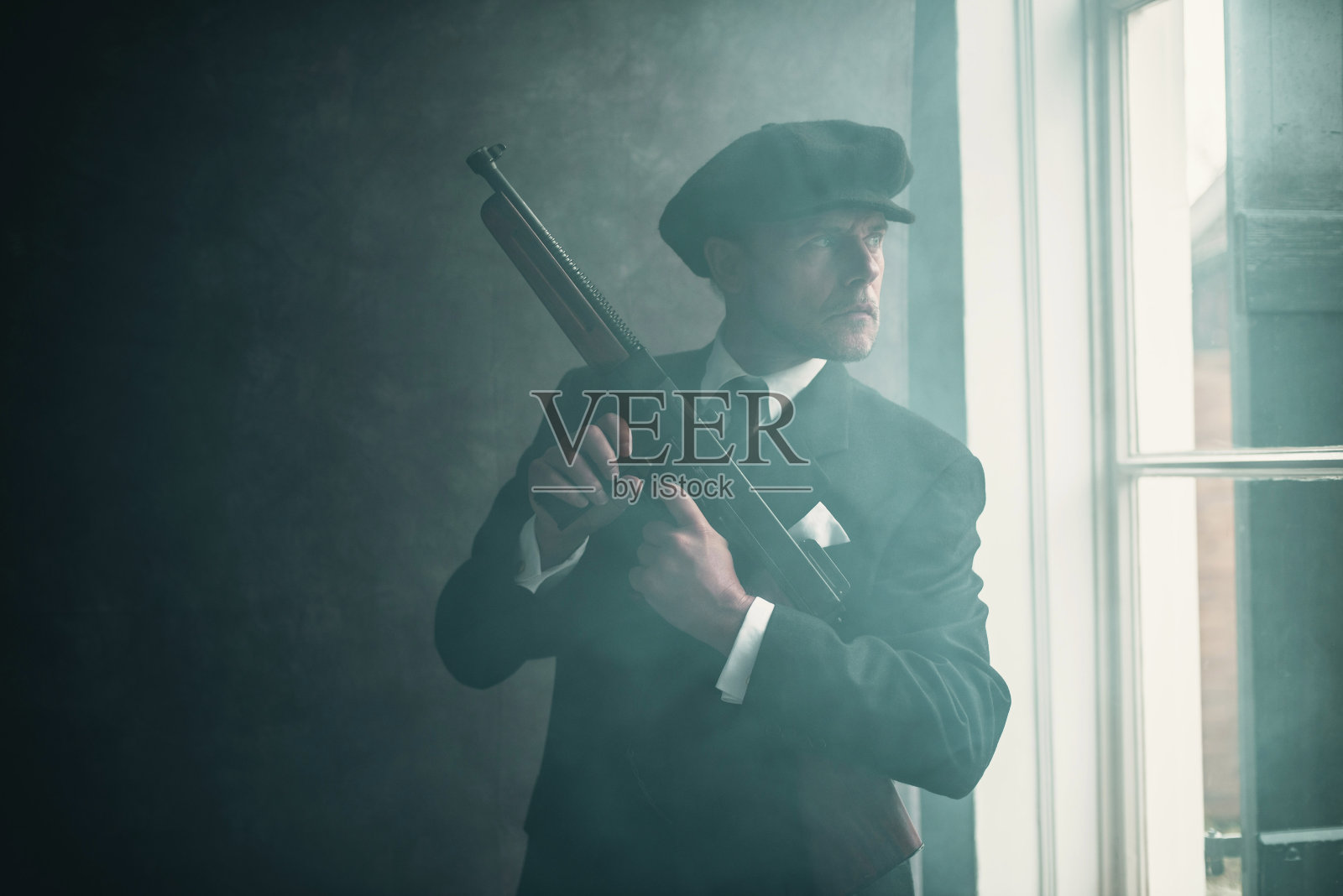 20世纪20年代复古的英国黑帮，头戴平帽，身穿西装。拿着枪站在那里望着窗外。照片摄影图片