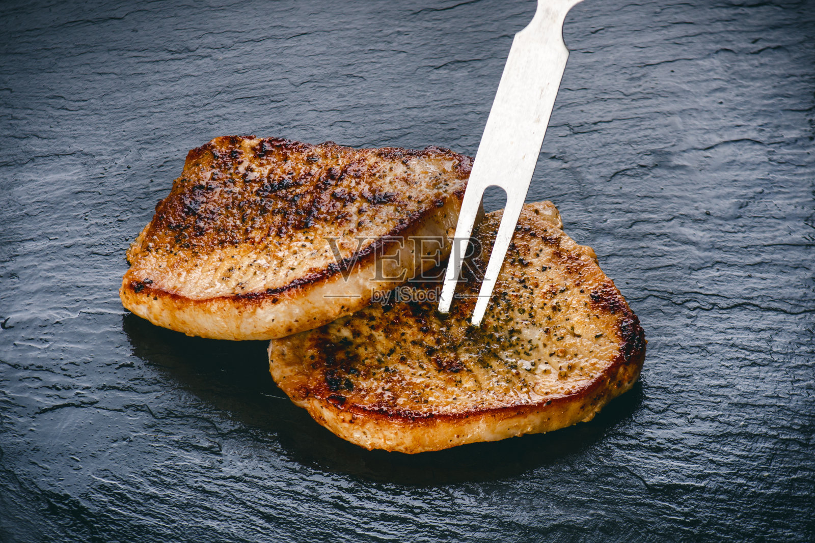 烤猪排，用叉子放在黑石板上的牛排。鲜肉和泡沫。黑暗的背景。烧烤和烧烤概念。照片摄影图片