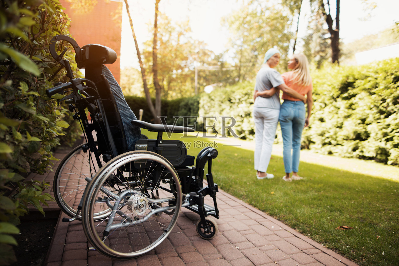 一位身患癌症的妇女和她的女儿站在一起。他们拥抱。旁边是一位坐轮椅的妇女。照片摄影图片