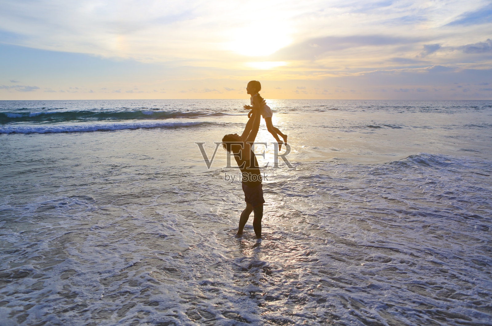 日落时分，父亲把小女孩扔在海滩上的剪影。照片摄影图片