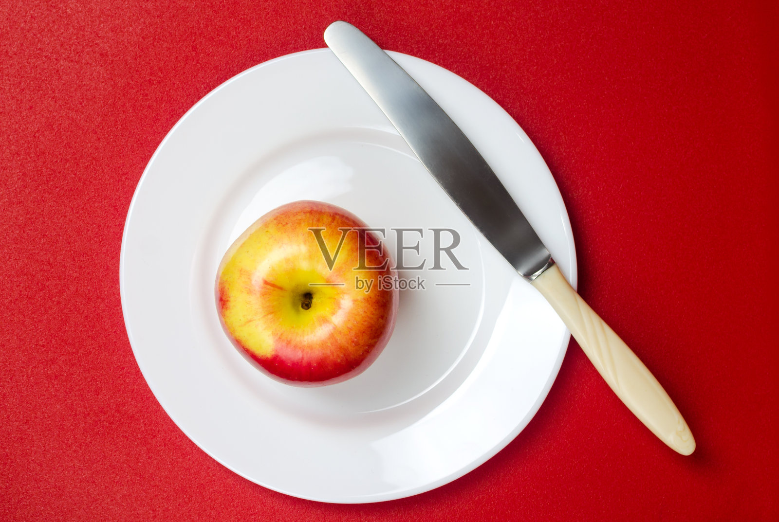 红色背景下的白色盘子上，用小刀切着成熟的红黄苹果照片摄影图片