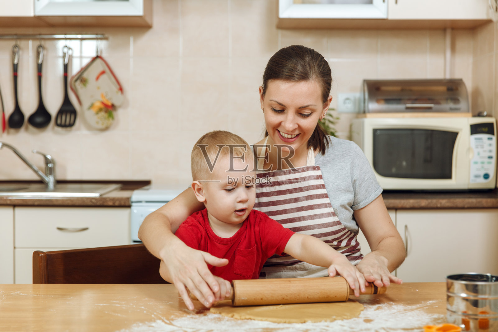 小男孩在灯光厨房里帮助妈妈做圣诞姜饼干。幸福的家庭妈妈30-35岁和孩子2-3在家里擀面团和剪饼干。关系和爱的概念照片摄影图片