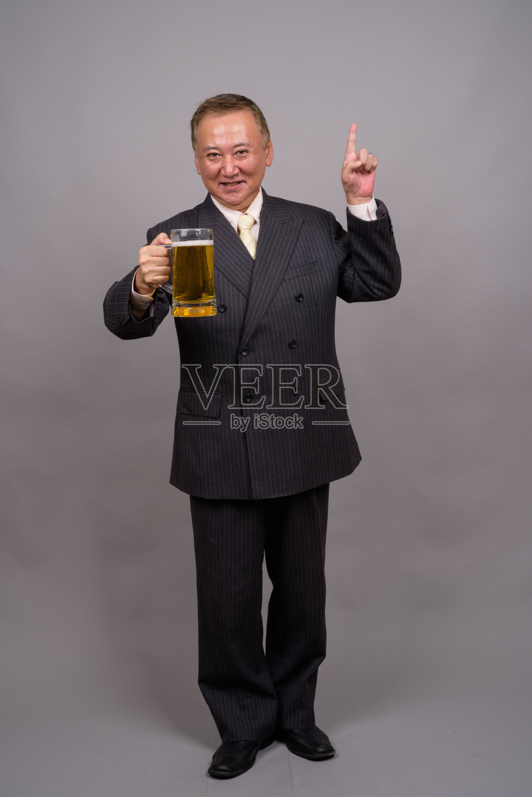 在灰色的背景下，一个成熟的亚洲商人拿着一杯啤酒照片摄影图片