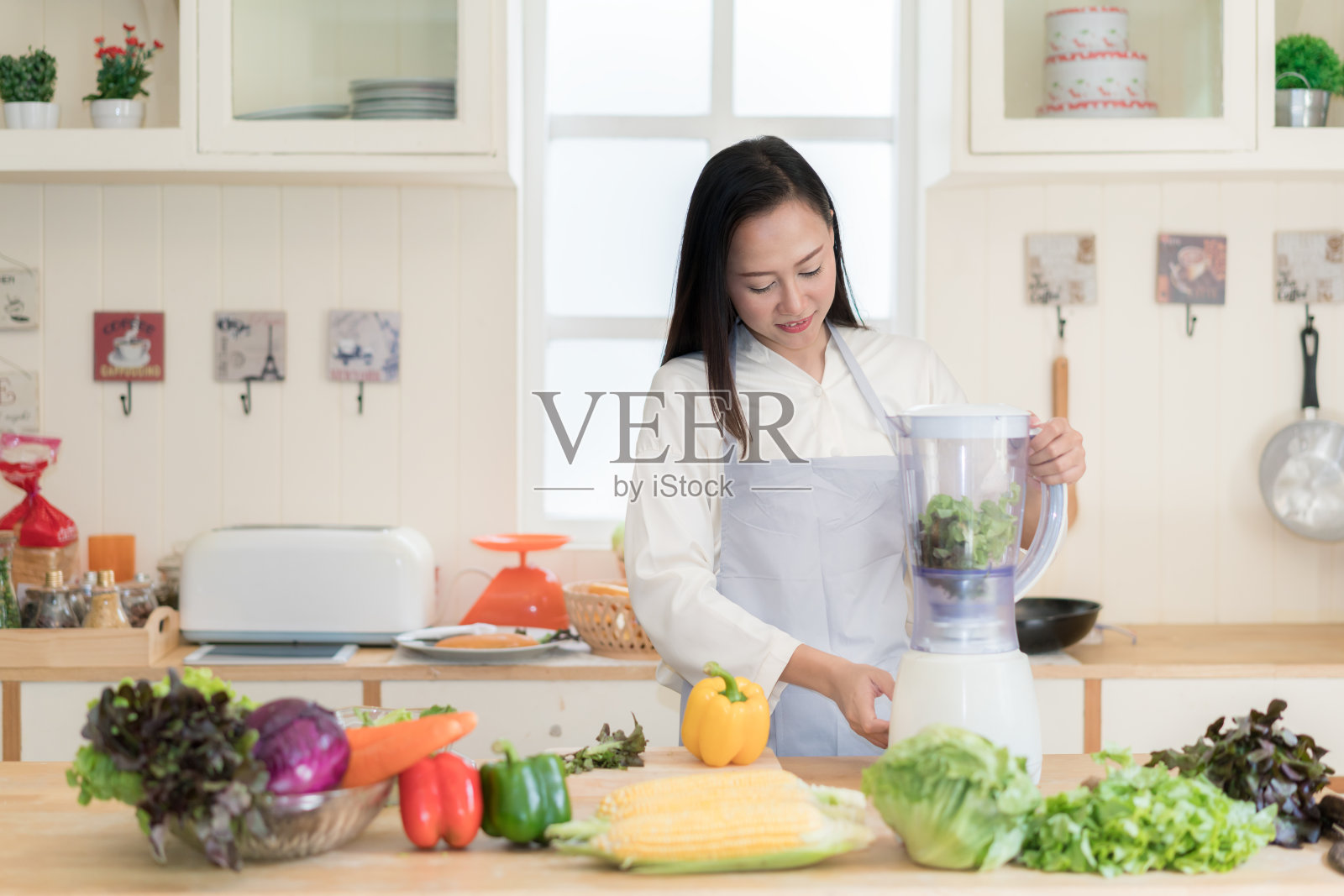 蔬菜奶昔。亚洲妇女在厨房用搅拌机做绿色冰沙。健康生食生活方式概念肖像美丽的年轻女子准备饮料与菠菜，胡萝卜，芹菜等。照片摄影图片