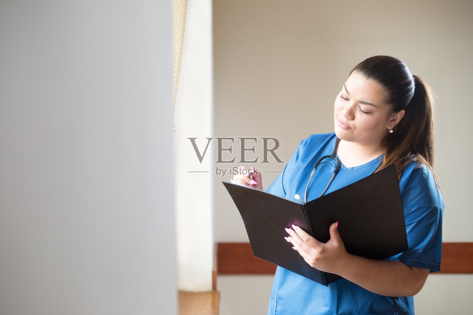 一位年轻的西班牙女医生正在分析一个临床病例，她手里拿着一个文件夹。一个女人穿着蓝色的制服，站在医院的窗户旁边。照片摄影图片