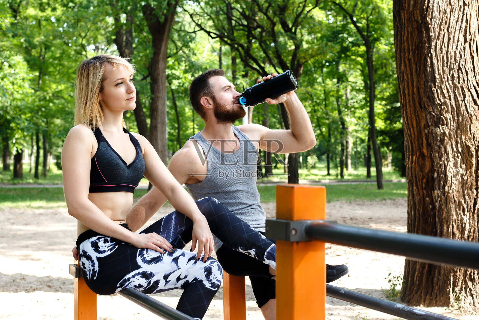 漂亮的金发女孩和留胡子的男人在户外公园锻炼后休息。一个人在喝黑瓶子里的水。照片摄影图片