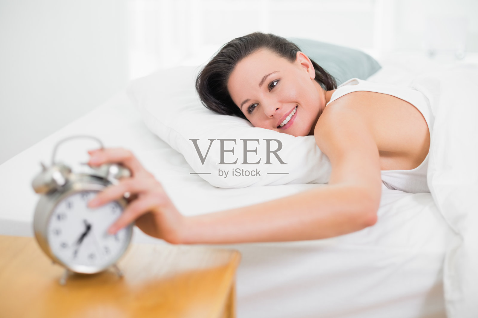 在床上微笑的女人向闹钟伸出手照片摄影图片