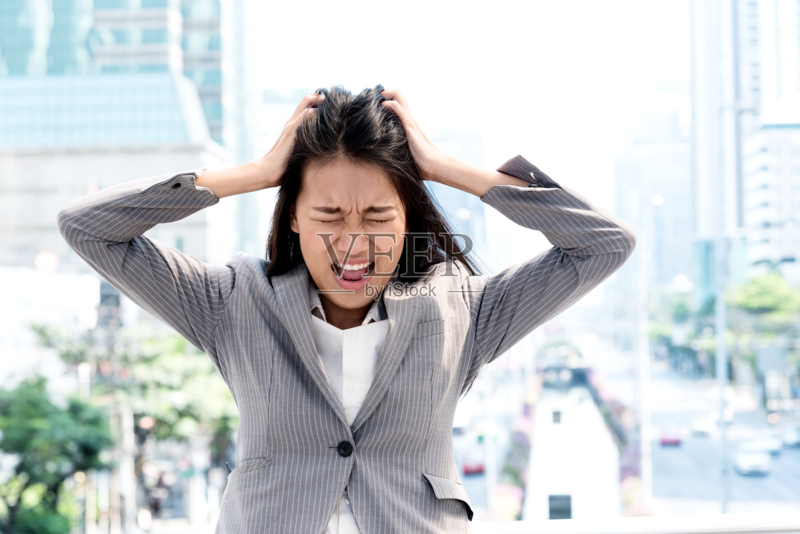 一个压力重重的年轻女商人双手抱头站在户外的肖像，背景是城市景观。不快乐的亚洲女孩与担心紧张的脸表情向下看。照片摄影图片